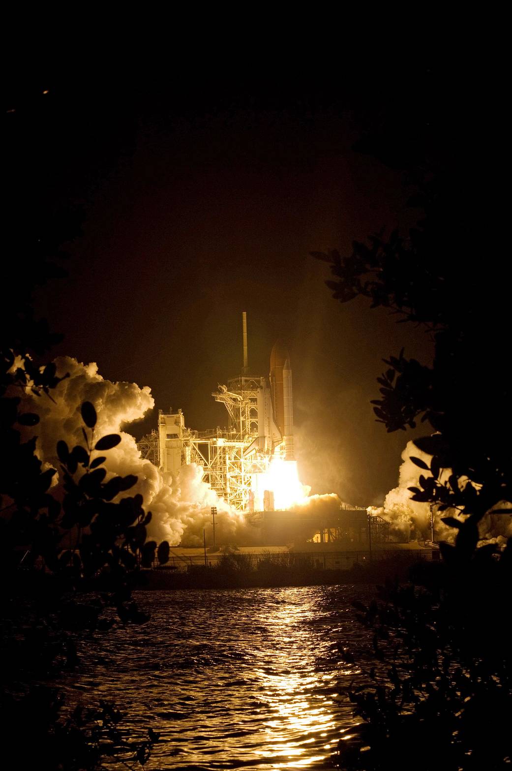 Fiery nighttime launch of shuttle Endeavour with foliage in the foreground at side of frame