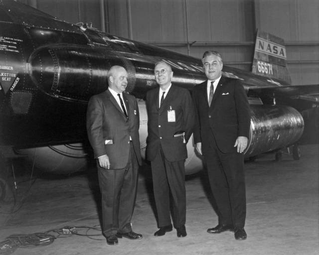 Three people in suits standing in front of the X-15 hypersonic aircraft.