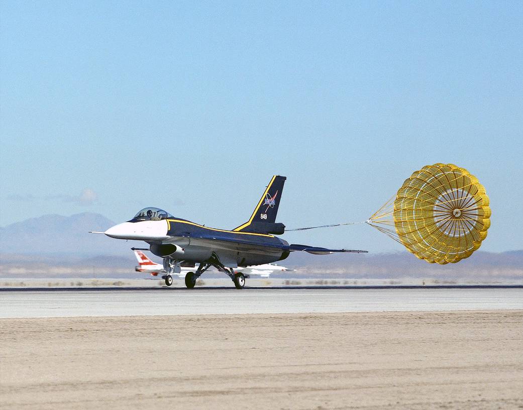 F-16XL aircraft landing, dragging a yellow parachute behind it.