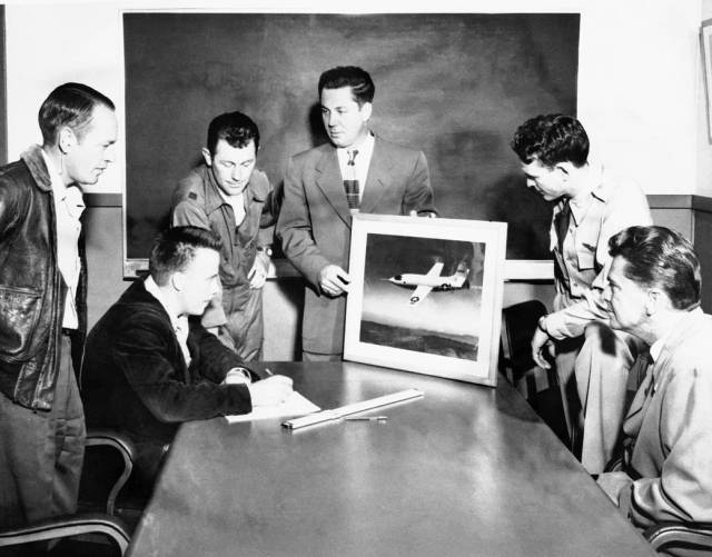Six people stand and sit around the end of a table looking at a picture of the X-1 aircraft. The person holding the picture is looking at another person.