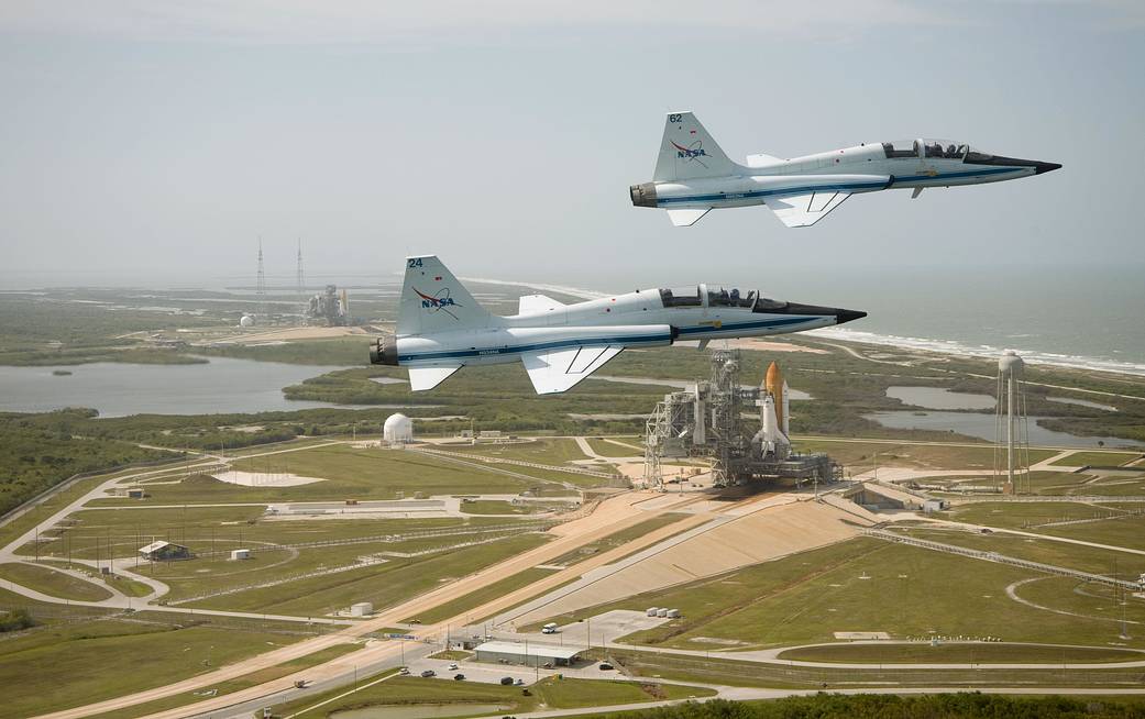 Two jets fly over launch pad with space shuttles vertical on pad below