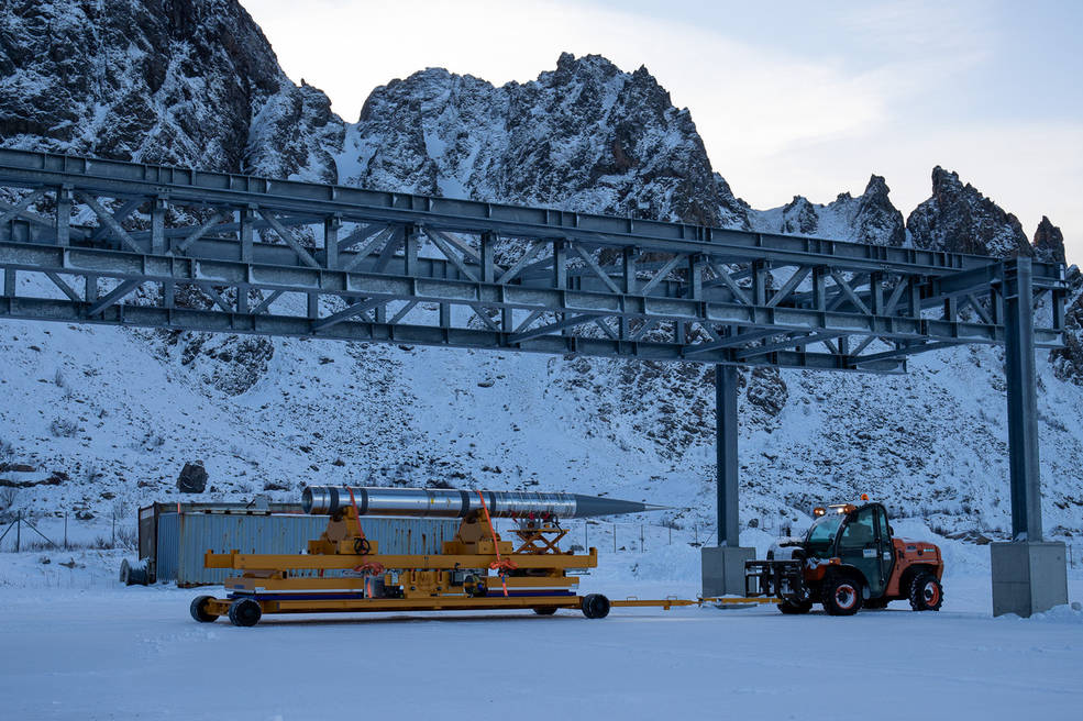 A snowy scene with mountains in the background. In the foreground, a part of a rocket is pulled by a small snowmobile tractor. Scaffolding made up of steel beams hang overhead.
