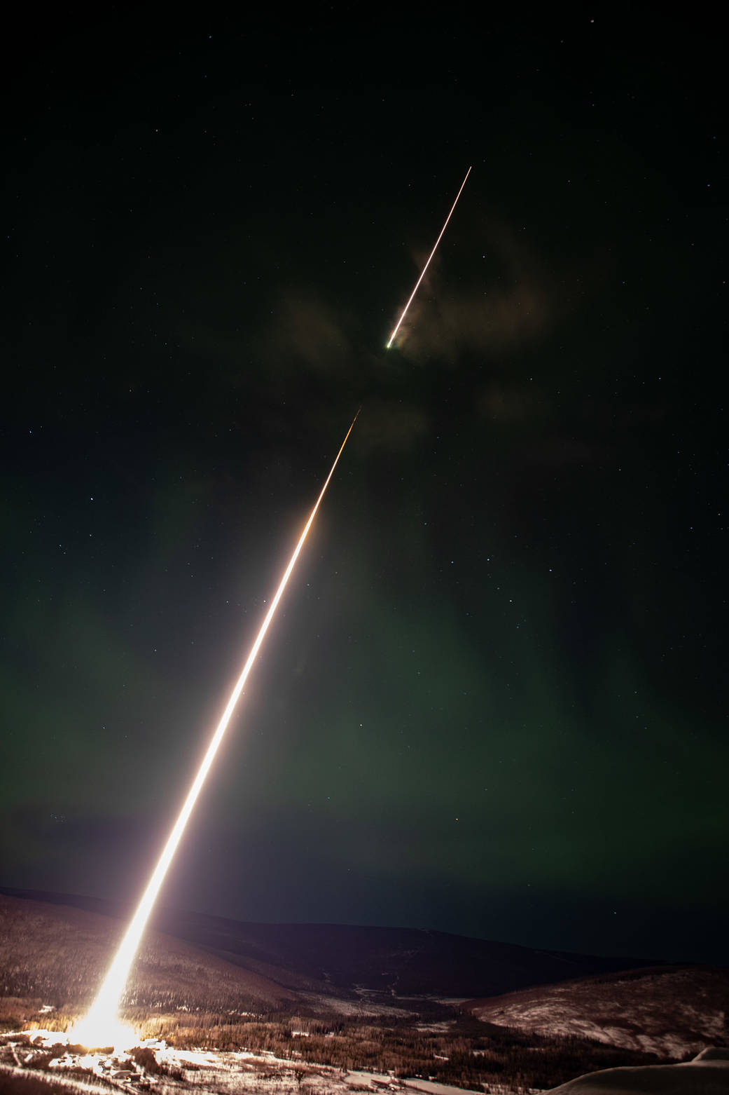 A long exposure photograph of a rocket launching into the sky. Long white streak from bottom left to top right, black sky and faint green aurora.
