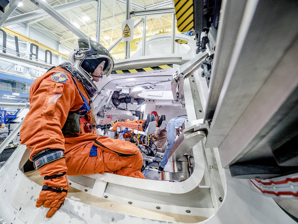 Inside a large room at Johnson Space Center, man in flight suit exits door of spacecraft