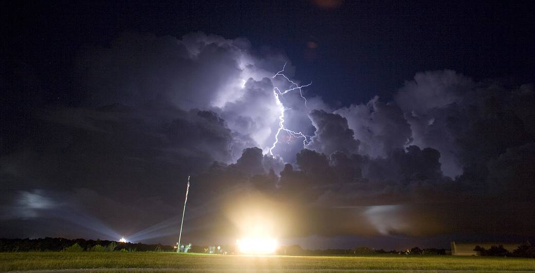 Bolts of lightning in dark sky at night with clouds over launch pad illuminated with lights