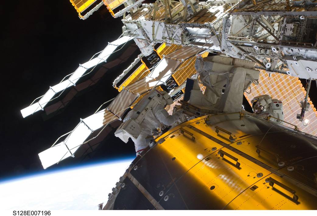 Bright gold cylindrical space station module with spacewalking astronauts in spacesuits on either side and solar arrays behind