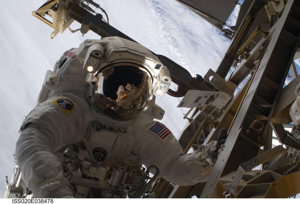 Spacewalking astronaut with Earth in background and reflection of other astronaut and solar arrays in astronaut's visor