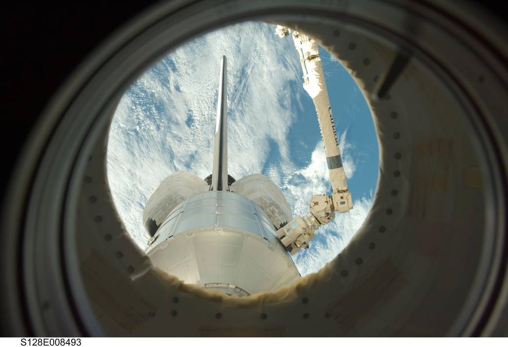 View through circular window at tail end of shuttle Discovery in orbit with Earth below
