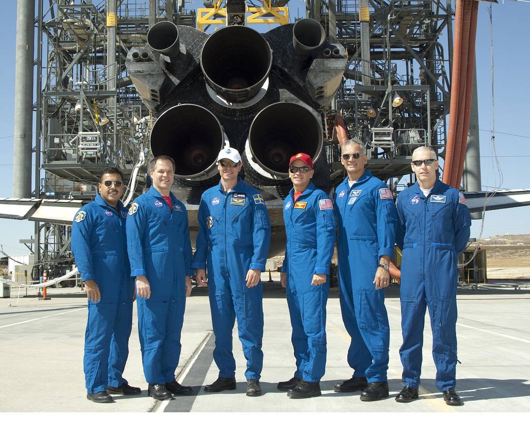 On runway, six astronauts pose for group photo wearing blue flight suits in front of the tail of shuttle Discovery