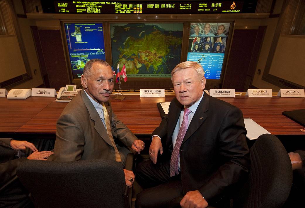 NASA Administrator Bolden seated at left and Roscosmos administrator seated at right inside mission control center, looking back