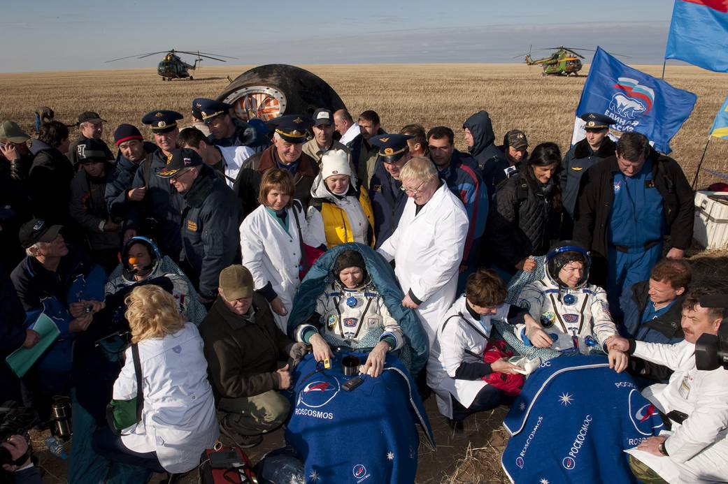 Three astronauts in spacesuits seated with blankets surrounded by people in field with spacecraft in background