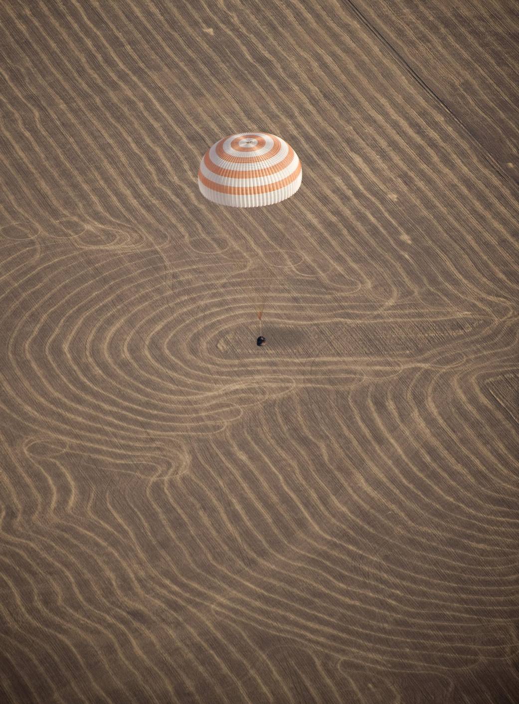 Spacecraft capsule descends toward field with red and white parachute above
