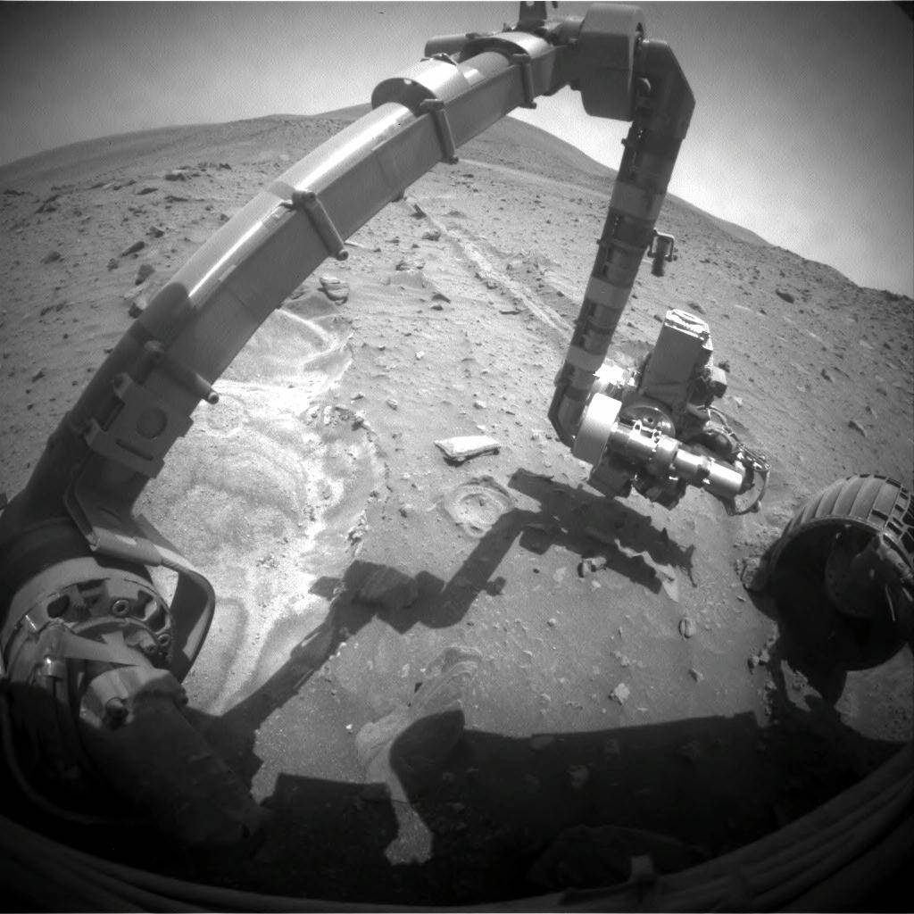 Rover's robotic arm reaches outward with shadow of rover below and Mars rocky terrain ahead