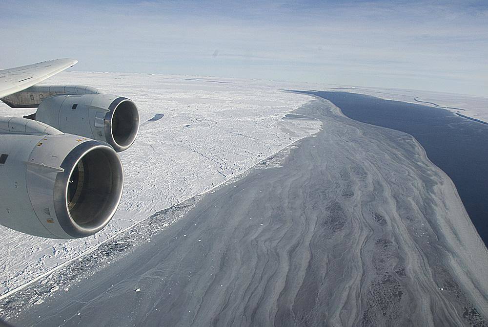 View from aircraft window over arc of large glacier sweeping over land below with aircraft engine visible at left