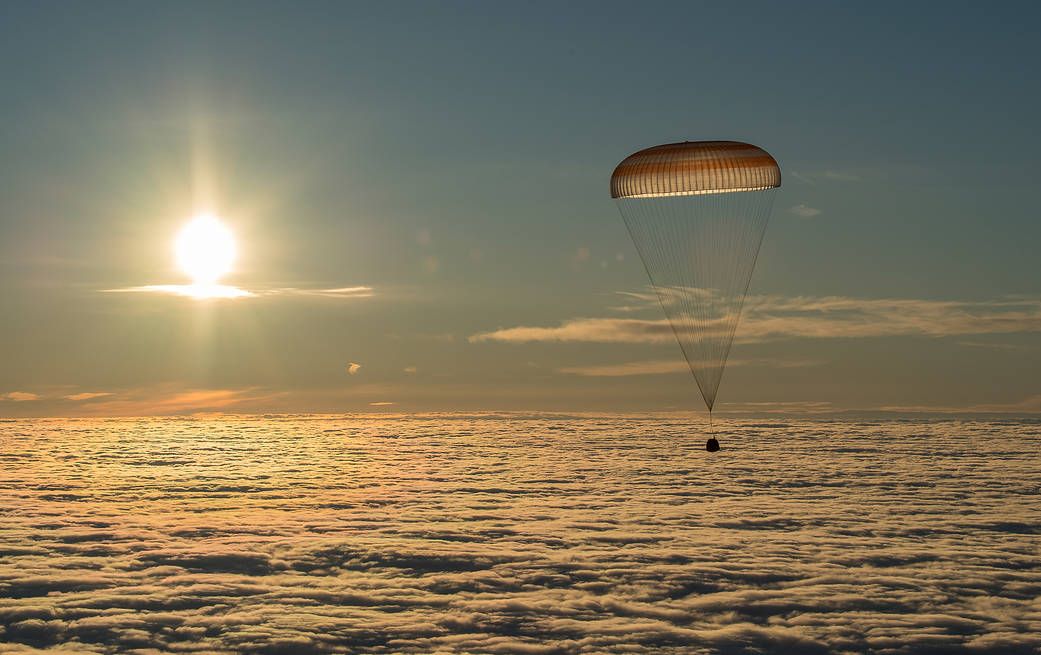 Soyuz with parachute deployed descends toward Earth with Sun in background