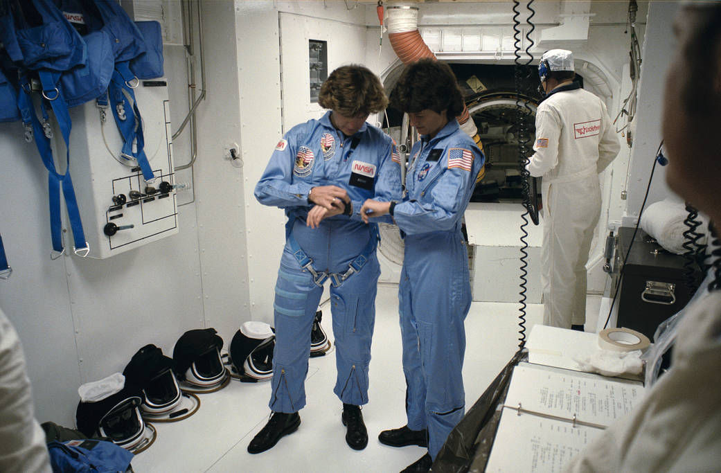 Astronauts Sally Ride and Kathryn Sullivan looking down at their watches inside room at Kennedy Space Center prior to launch