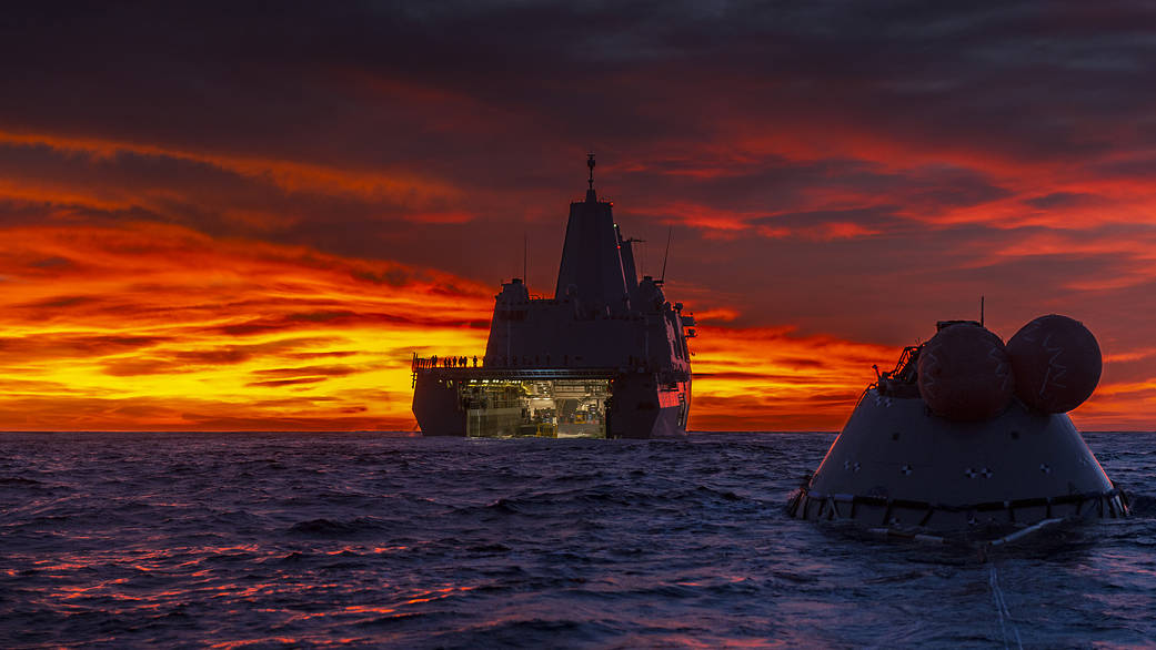 Brilliant red and yellow sunrise over the ocean with the Orion test capsule in front of the USS Murtha ship 