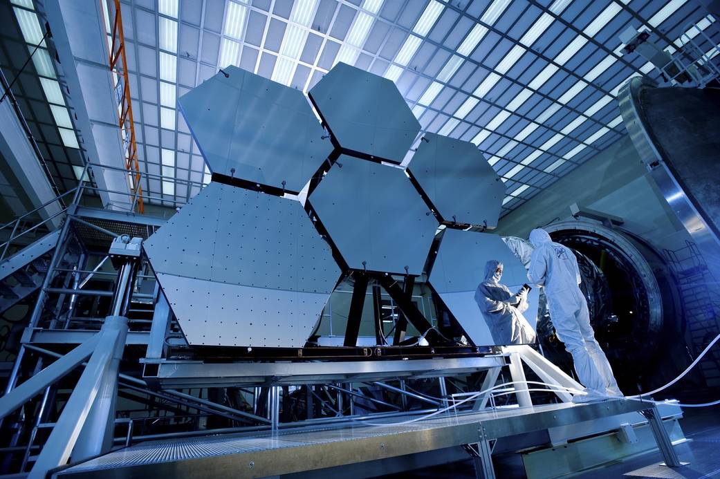 Six hexagonal large mirrors for Webb Telescope arranged three above three with technician in clean suit reflected in lower right