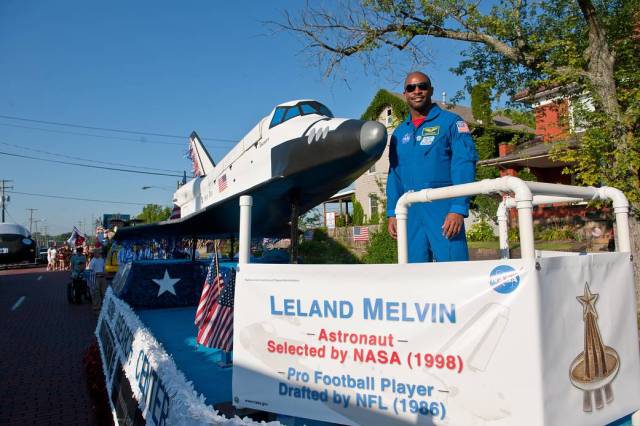 Astronaut Leland Melvin in blue flight suit wearing sunglasses next to shuttle model on parade float