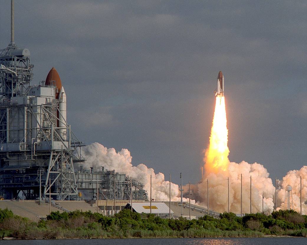 In the distance, shuttle Discovery launches in daytime, with shuttle Columbia on launch pad visible at far left of image