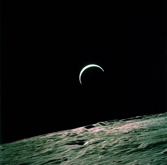The Earth appears in the black sky as a slim crescent of white-blue. In the foreground is the rough, rocky surface of the Moon.