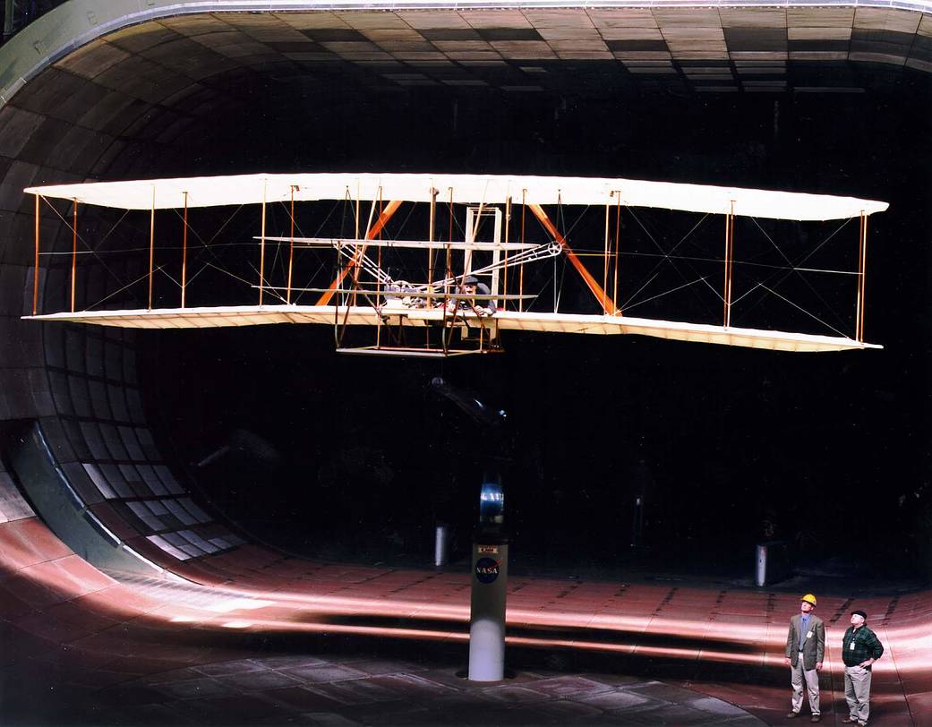 Model of Wright brothers' aircraft in large wind tunnel and two technicians standing below looking up toward it