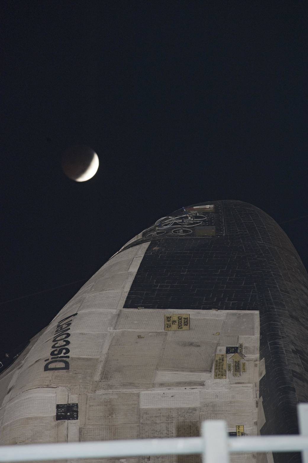 Nighttime closeup of nose of shuttle Discovery with crescent of moon during eclipse above