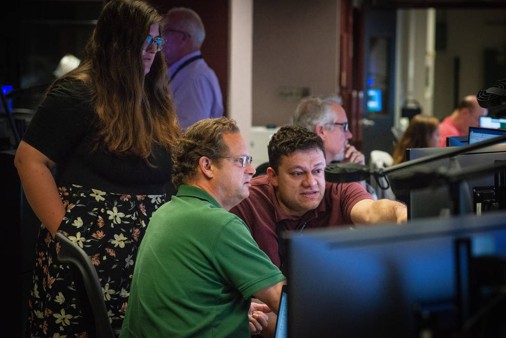 A group of people sits at a long table with many computer monitors. Two men in the foreground point and look at a monitor facing away from the camera. Hannah Daelemans, a woman wearing a black tee and skirt, stands behind.   