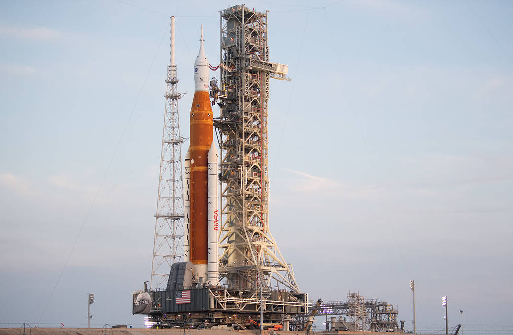 Artemis I sits atop the launch pad. The sky in the background is a pale blue, with a few streaks of white clouds. The SLS is a rich orange color.
