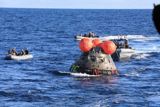 Grey cone-shaped spacecraft on the surface of the water, with three orange balloons at its top. There are three grey motor boats on its way to the spacecraft, with white foam in their wake.