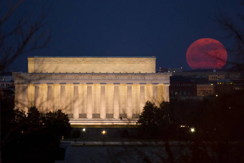 Supermoon appearing very large and deep red at upper right of image with Lincoln memorial building in foreground at left