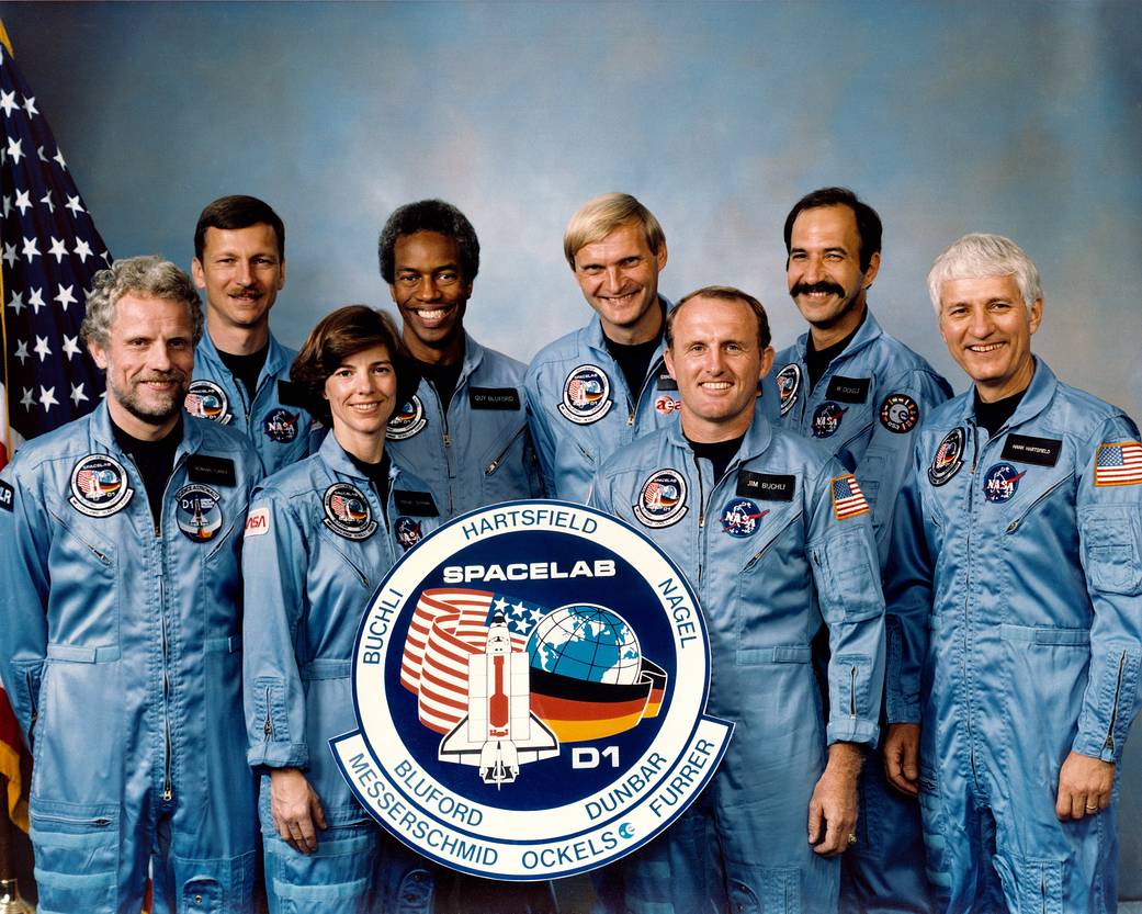 Eight astronauts stand and pose for their crew picture. They are wearing space flight suites and smiling.