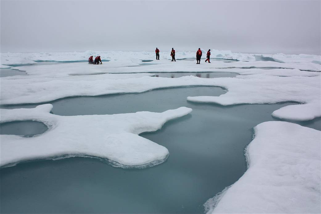 In the distance scientists walk on Arctic sea ice surrounded by ponds of melt water