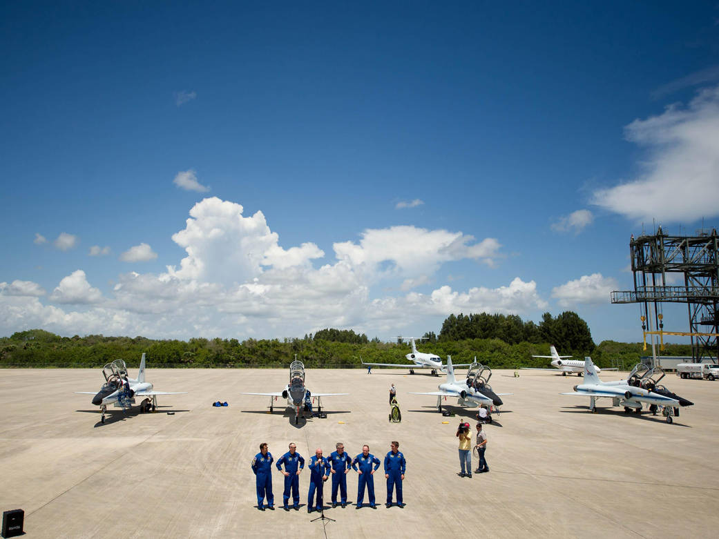 Six astronauts in flight suits on tarmac with jets parked behind and blue sky with clouds overhead
