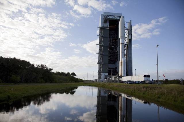 At Space Launch Complex 41, a pond reflects the 197-foot-tall United Launch Alliance Atlas V rocket and Vertical Integration Facility against a blue sky flecked with white clouds. 