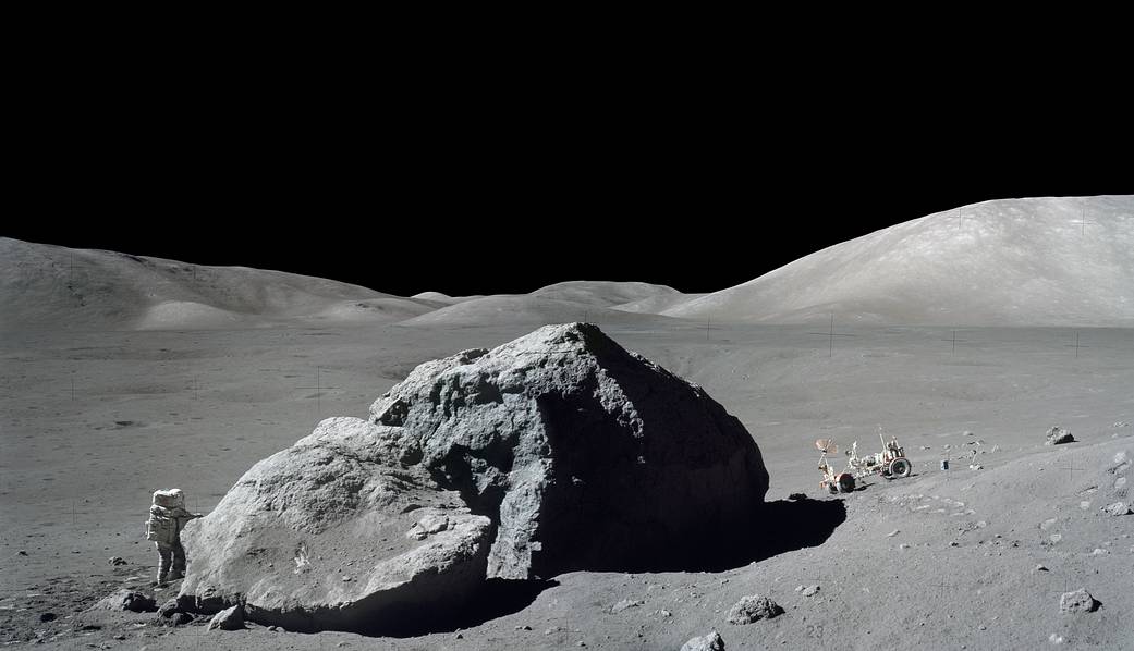 Astronaut on spacewalk on lunar surface near large rock with moon rover at right