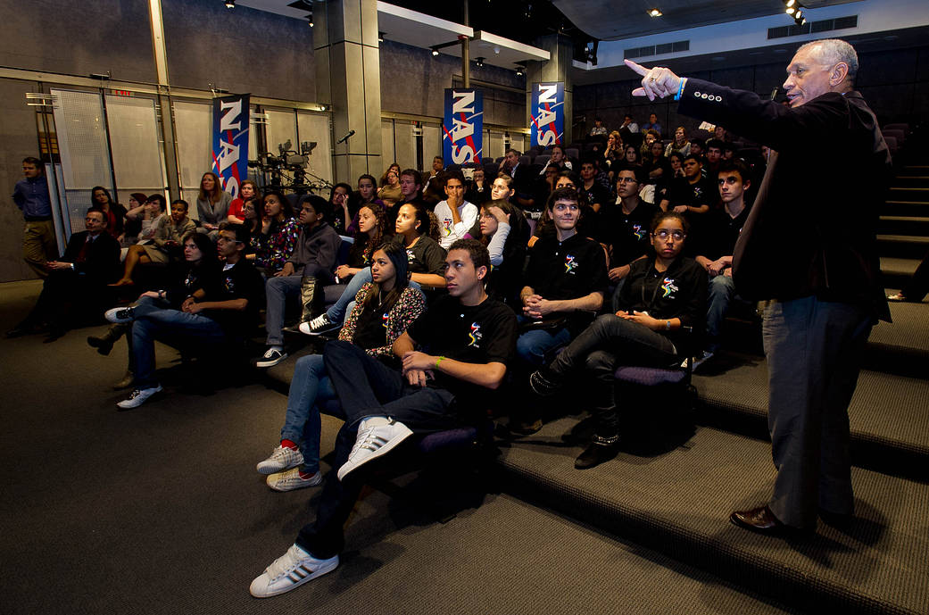 Large group of young adults in auditorium looking toward front as NASA Administrator Bolden standing at right points forward