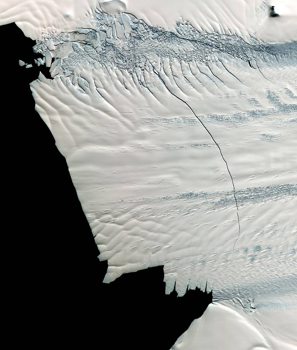 View from above of flat white sheet of glacier ice with ridges in ice and large crack in center with deep blue water at left