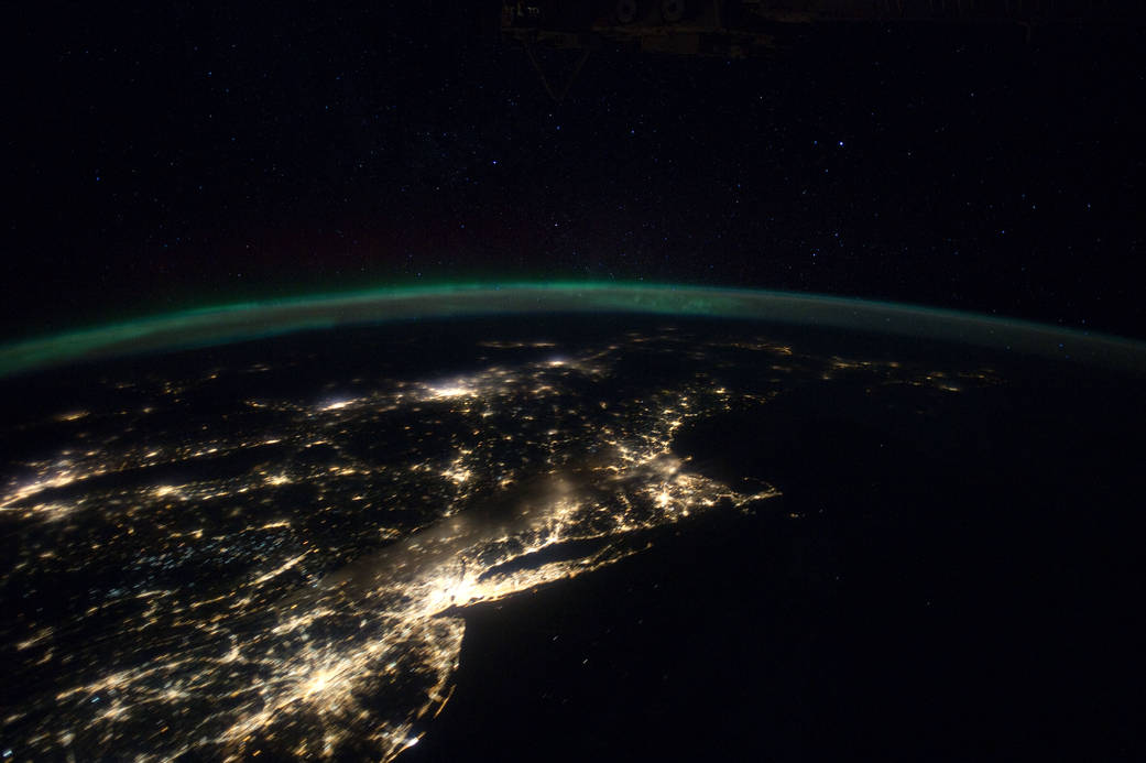 Arc of Earth's atmosphere in green runs across image horizontally in nighttime image with brightly lit cities below