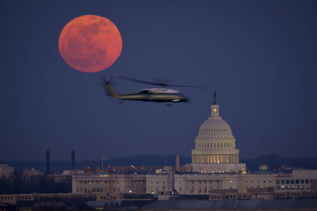 Large full moon appearing orange in night sky at left with Capitol building at bottom and helicopter flying past at center