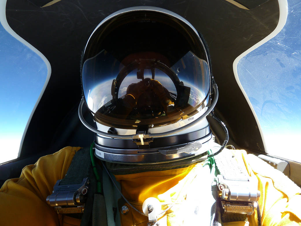 In-flight selfie of a pilot wearing a spacesuit. The pilot's closed helmet shows a reflection of what he sees in front of him.