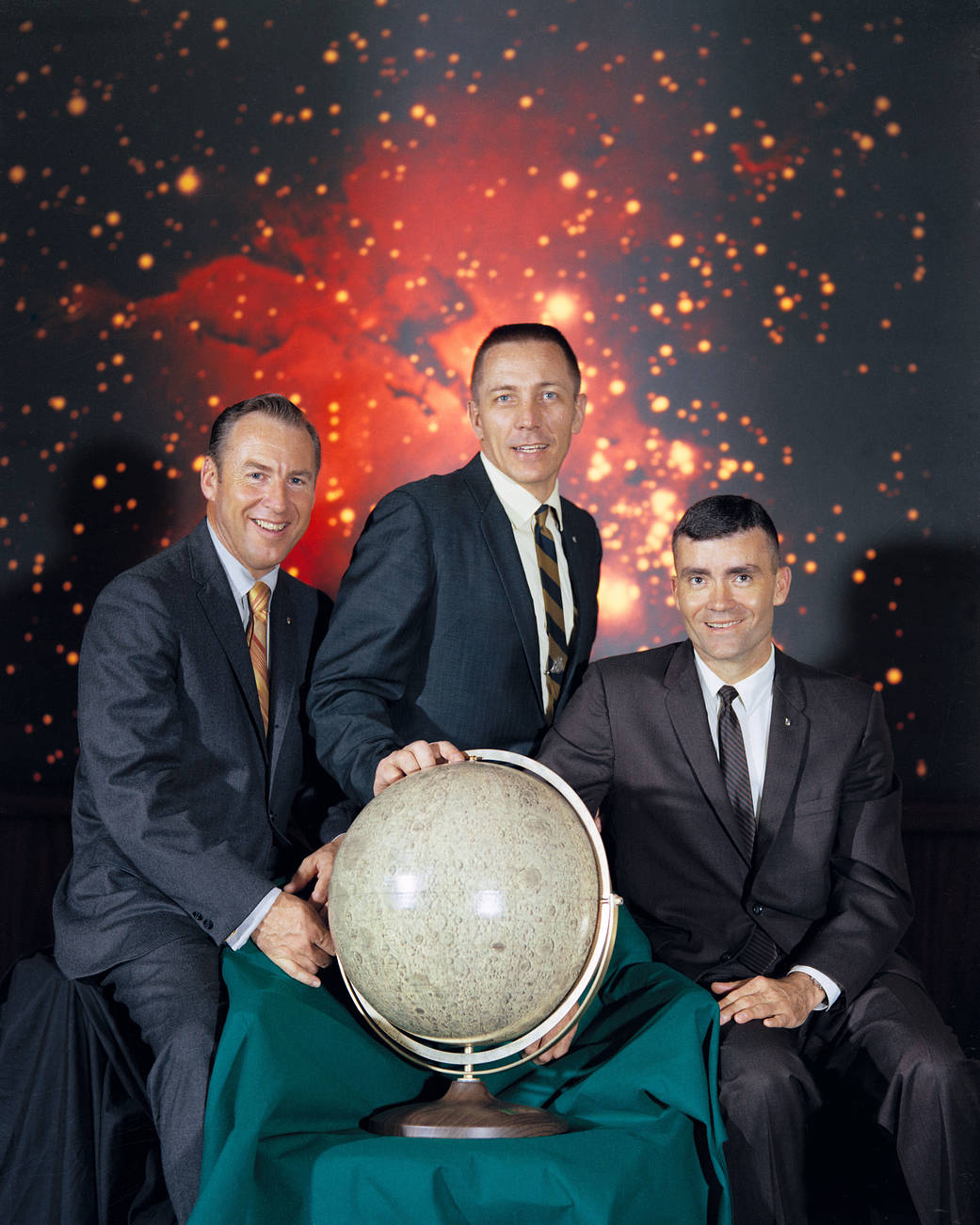 Apollo 13 crew pose for portrait wearing suits with globe of moon and galaxy backdrop