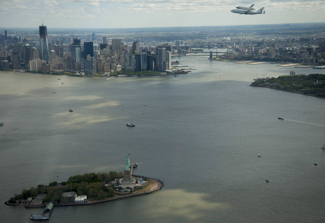 Skyline of New York City with Hudson river in front and shuttle Enterprise atop carrier aircraft flying in upper right