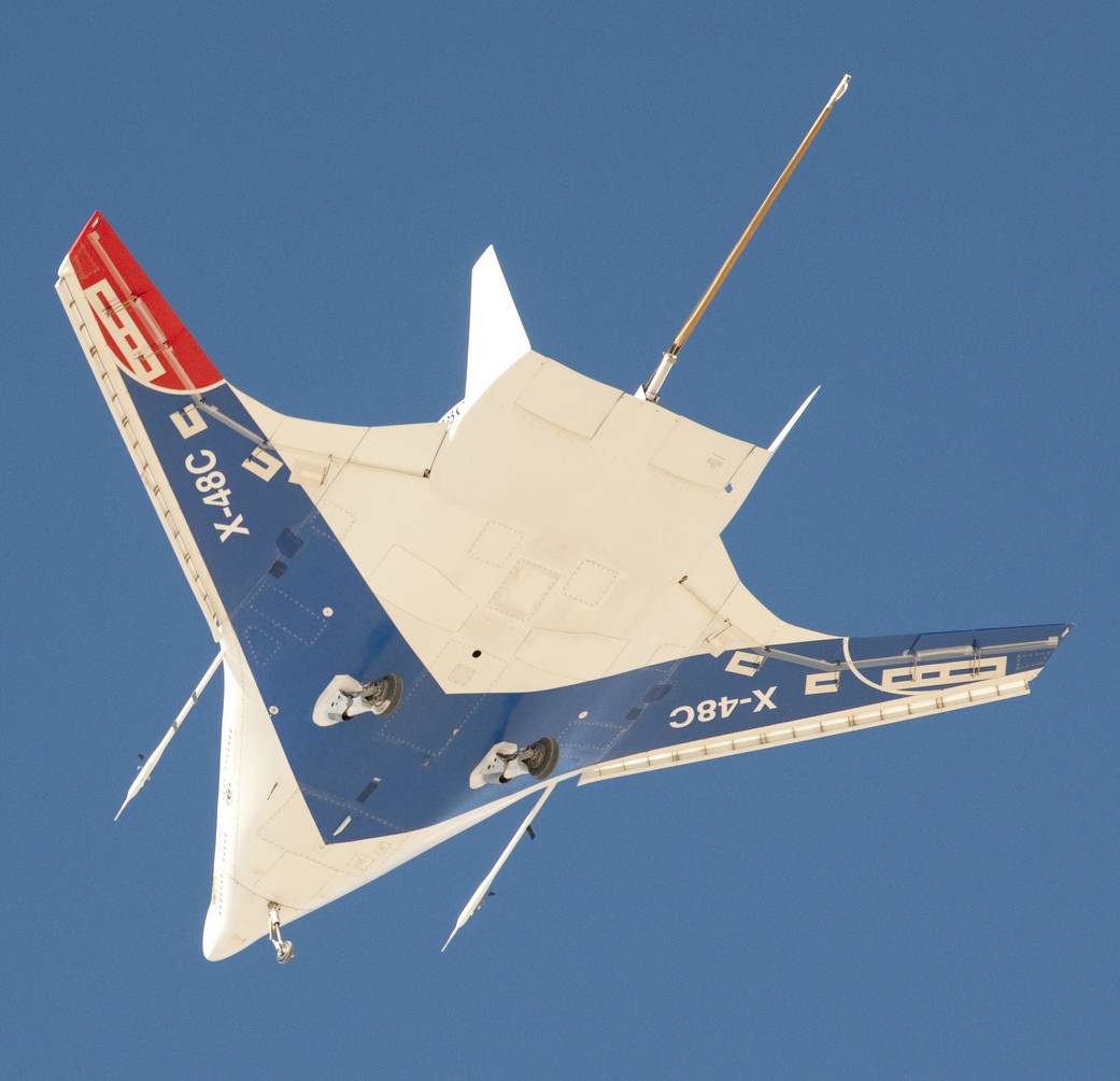 Image from on the ground of the blended wing body X-48C aircraft above in flight against blue skies.
