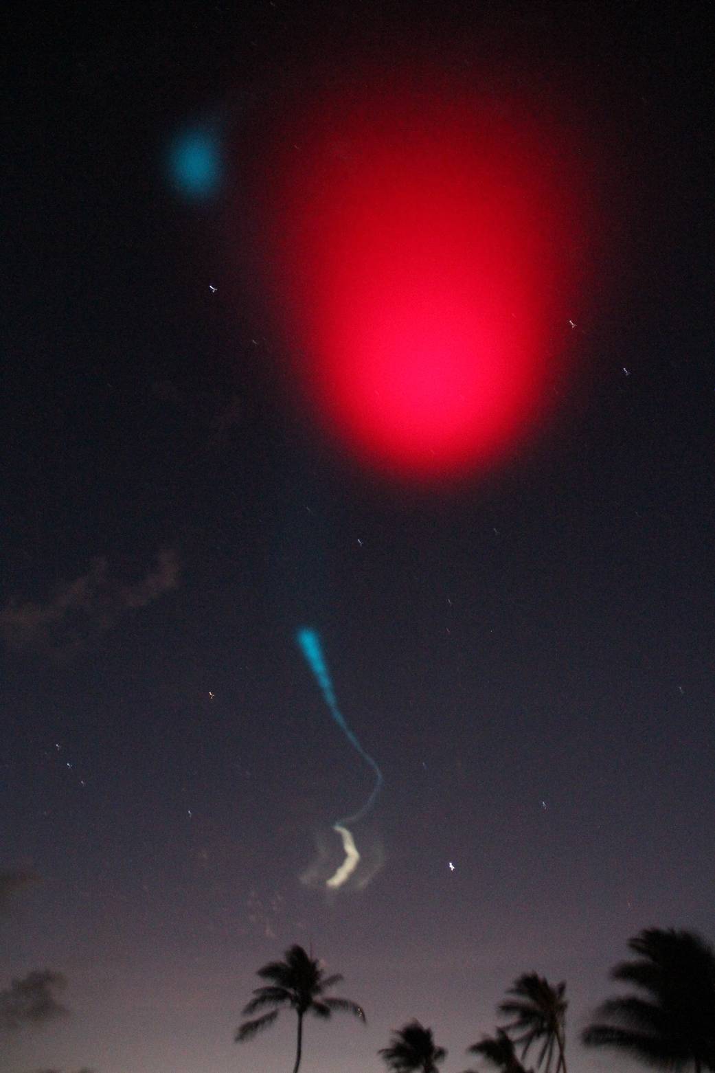 trails from nighttime rocket launch in night sky with palm trees below and bright red light above
