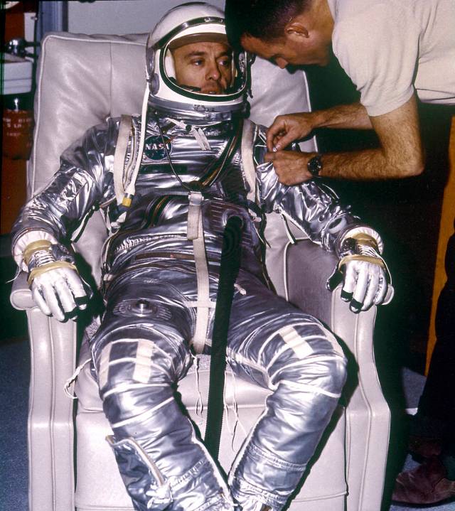 Astronaut Alan Shepard, seated, while technician checks his silver pressure suit