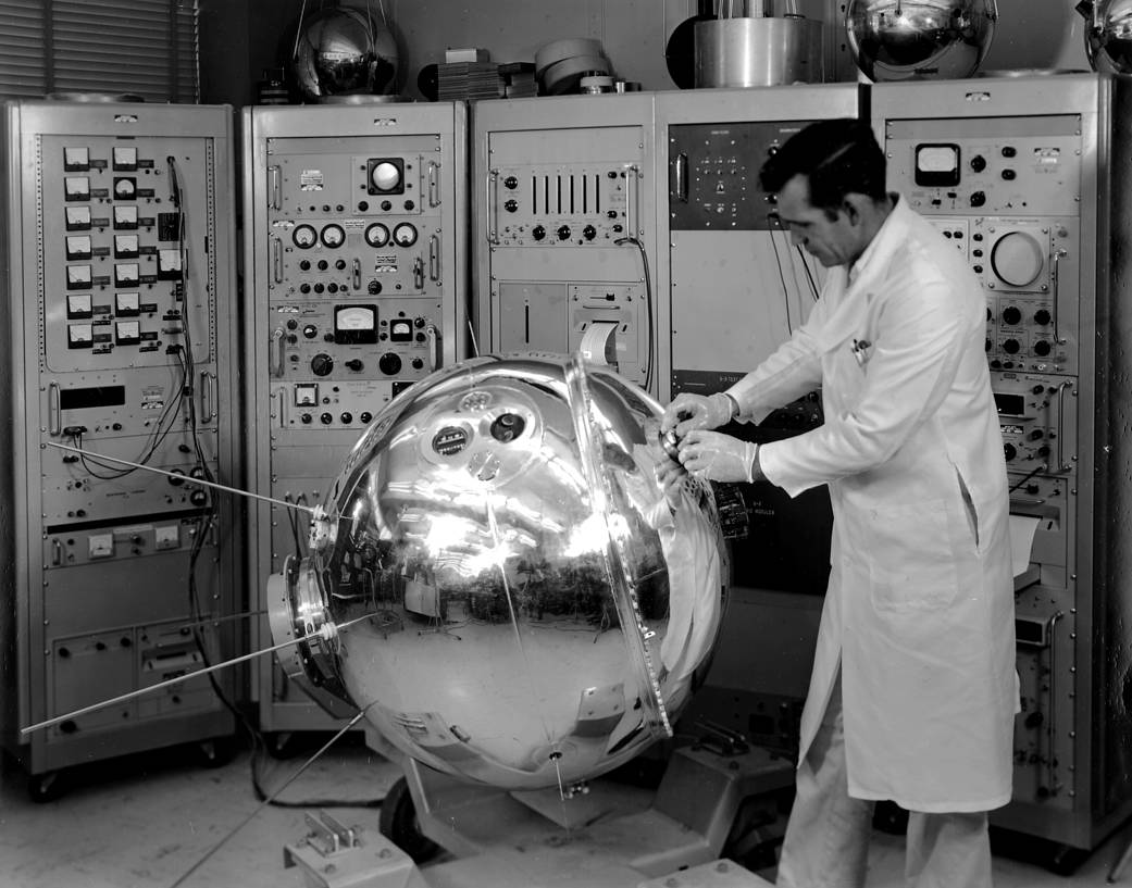 Technician examines silver round Explorer satellite on floor inside room at NASA Goddard