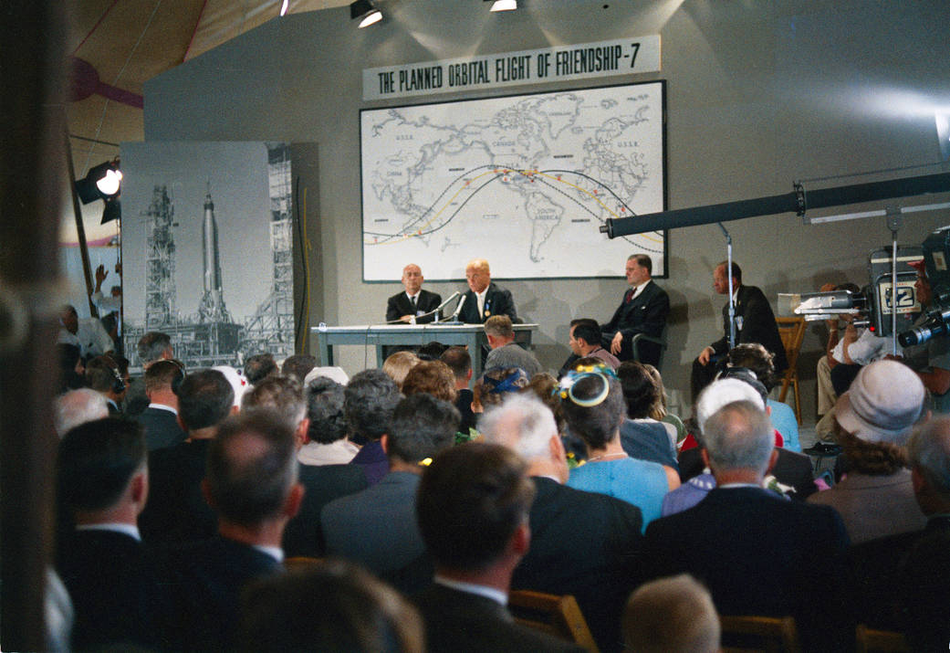 News conference for the Mercury-Atlas 6 mission with image of Friendship 7 spacecraft orbital trajectory in background.