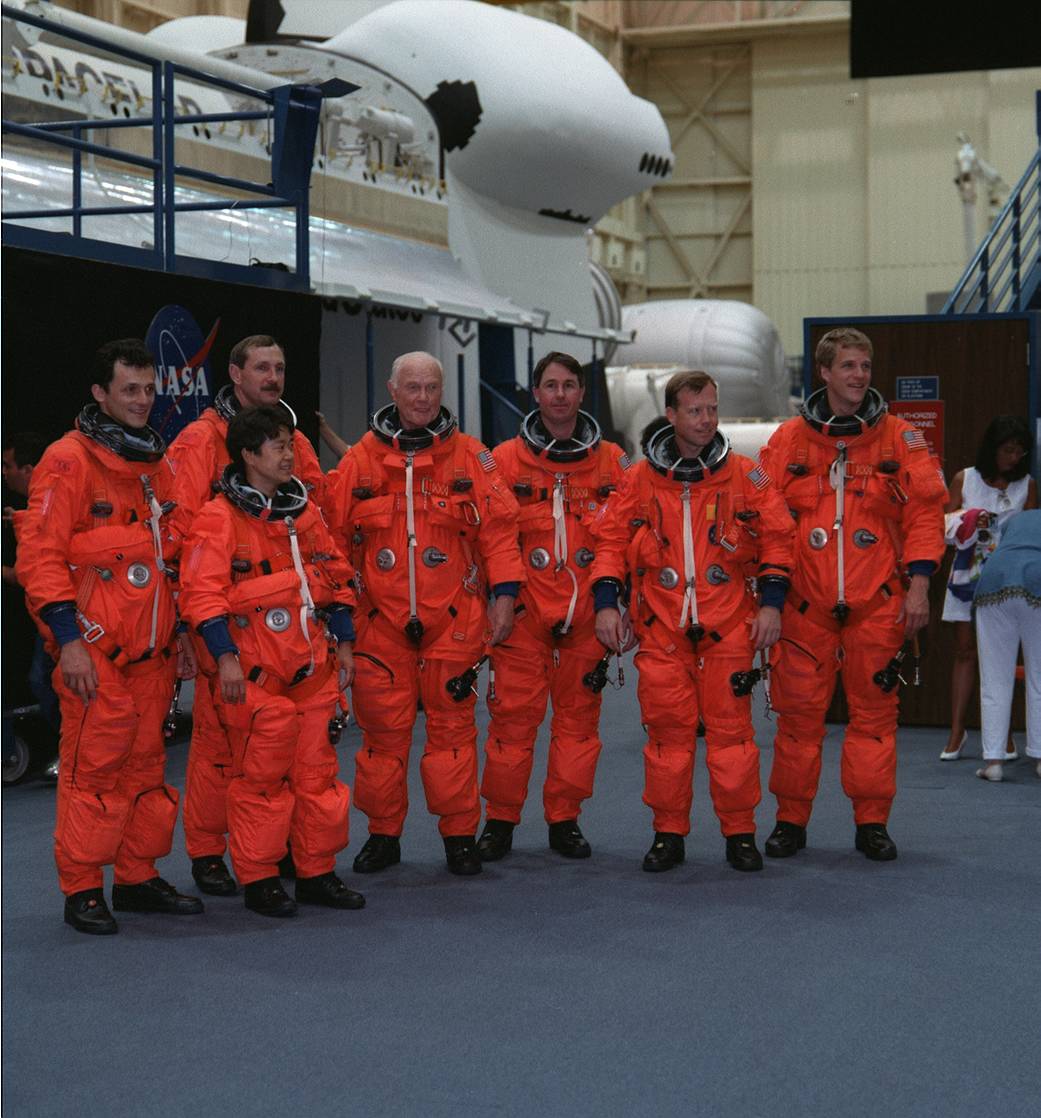 Seven astronauts wearing orange spacesuits without helmets pose for group photo in large training room
