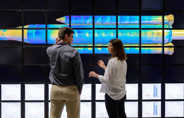 Two people standing front of a wall of screens showing a multicolored image of a rocket body